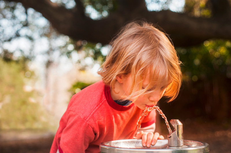 Junge trinkt Wasser an einem Trinkbrunnen. Im Hintergrund ist ein Park zu sehen.